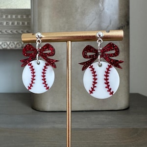 May include: A pair of baseball-themed earrings. Each earring features a white disc with red stitching, resembling a baseball, topped with a sparkly red bow. The earrings hang from silver-colored posts.