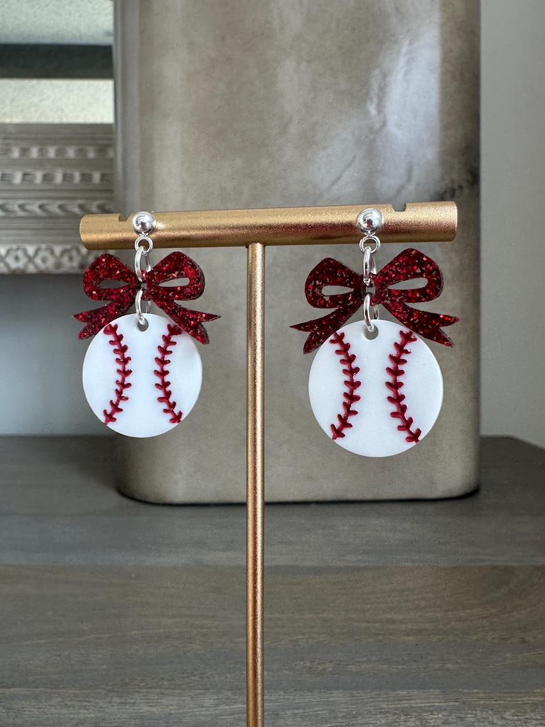 May include: A pair of baseball-themed earrings. Each earring features a white disc with red stitching, resembling a baseball, topped with a sparkly red bow. The earrings hang from silver-colored posts.