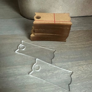 May include: A stack of brown paper gift tags with a hole for string or ribbon. Two clear plastic gift tags with a hole for string or ribbon are also shown.