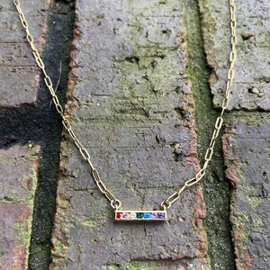 May include: A gold-colored necklace with a rectangular pendant featuring a row of colorful gemstones. The gemstones are red, clear, green, blue, and purple. The necklace chain has an elongated oval link design.