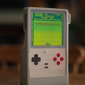Könnte beinhalten: Eine tragbare Spielkonsole mit einem hellgrauen rechteckigen Gehäuse. Der Bildschirm zeigt eine grüne Benutzeroberfläche mit dem Text LEVEL 01, SCORE 000000 und PUSH KEY. Sie verfügt über ein schwarzes Steuerkreuz, vier rote Tasten und zwei kleine graue Tasten.
