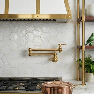 Op de afbeelding: Een keuken met een koperen pot op een zwart en goud fornuis. Een gouden kraan is gemonteerd op een witte zeshoekige tegelachterwand. Een witte afzuigkap met gouden afwerking bevindt zich boven het fornuis. Plankjes bevatten witte kommen, glazen en een groen vogelbeeldje.