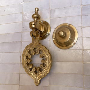 Puede incluir: Aldaba de puerta de lat&oacute;n dorado con un dise&ntilde;o ornamental calado. El juego incluye una placa de golpe a juego. El conjunto est&aacute; montado en una pared de azulejos blancos. La aldaba tiene una est&eacute;tica vintage.