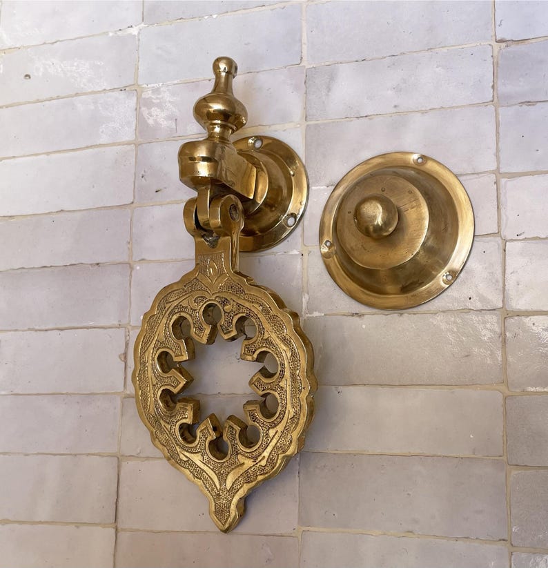 Puede incluir: Aldaba de puerta de lat&oacute;n dorado con un dise&ntilde;o ornamental calado. El juego incluye una placa de golpe a juego. El conjunto est&aacute; montado en una pared de azulejos blancos. La aldaba tiene una est&eacute;tica vintage.