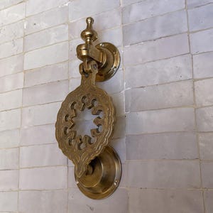 Puede incluir: Aldaba de puerta de lat&oacute;n ornamentada con un dise&ntilde;o calado decorativo. La aldaba presenta una placa circular y un asa en forma de l&aacute;grima, montada en una pared de azulejos rectangulares blancos. El lat&oacute;n tiene un tono dorado c&aacute;lido.