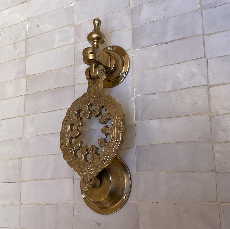 Puede incluir: Aldaba de puerta de lat&oacute;n ornamentada con un dise&ntilde;o calado decorativo. La aldaba presenta una placa circular y un asa en forma de l&aacute;grima, montada en una pared de azulejos rectangulares blancos. El lat&oacute;n tiene un tono dorado c&aacute;lido.
