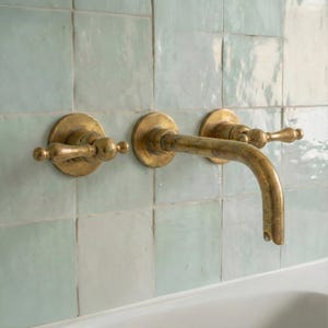 May include: A brass faucet with two handles and a curved spout, mounted on a light blue tiled wall. The faucet's design is vintage-inspired, with decorative handles. The tiles are square and have a subtle, irregular texture.