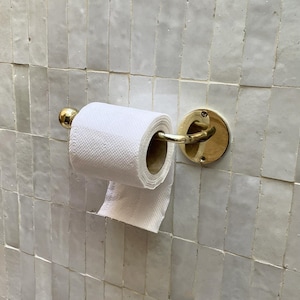 May include: A gold-toned metal toilet paper holder with a roll of white toilet paper. The holder is mounted on a white tiled wall. The holder has a circular base and a curved arm to hold the paper roll.
