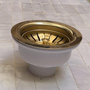 Puede incluir: Un colador de fregadero de cocina de color latón con un cuerpo blanco. El colador tiene un diseño circular con una serie de ranuras y un botón central. El borde es de color dorado brillante y el cuerpo es blanco.