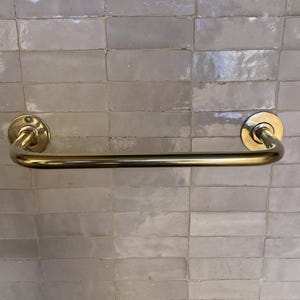 May include: A gold-colored towel bar with a curved design. The bar is mounted on a light gray tiled wall. The bar has a circular mounting plate on each end.