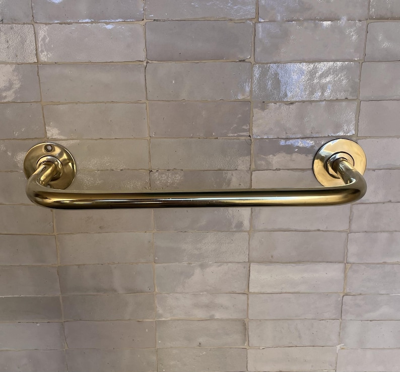 May include: A gold-colored towel bar with a curved design. The bar is mounted on a light gray tiled wall. The bar has a circular mounting plate on each end.