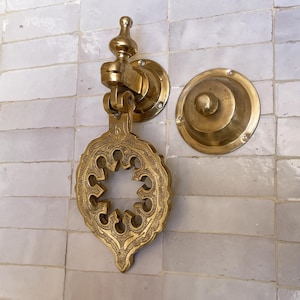 Puede incluir: Aldaba de puerta de lat&oacute;n dorado con un dise&ntilde;o ornamental calado. Est&aacute; montada sobre una superficie de azulejos blancos. Tambi&eacute;n hay una placa de golpe redonda a juego. La aldaba tiene una est&eacute;tica vintage detallada.
