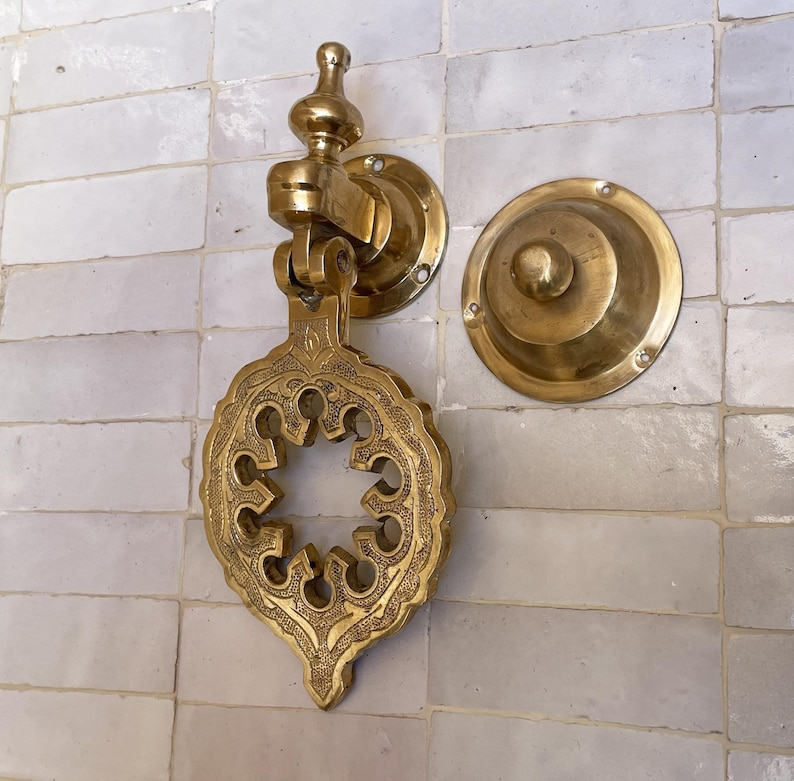 Puede incluir: Aldaba de puerta de lat&oacute;n dorado con un dise&ntilde;o ornamental calado. Est&aacute; montada sobre una superficie de azulejos blancos. Tambi&eacute;n hay una placa de golpe redonda a juego. La aldaba tiene una est&eacute;tica vintage detallada.