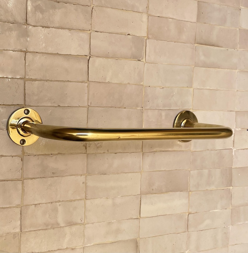 May include: A gold-colored grab bar, mounted on a light-colored tiled wall. The bar is a simple, curved design with circular mounting plates on each end. The wall tiles are rectangular and have a slightly uneven surface.