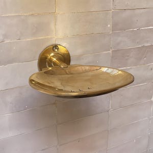 Pode incluir: Um saboneteira de latão montada na parede com uma placa traseira circular e um prato curvo em forma oval. O prato é montado em uma parede de azulejos retangulares de cor clara. O latão tem um acabamento brilhante e reflexivo.