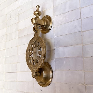Puede incluir: Aldaba de puerta de lat&oacute;n dorado con un dise&ntilde;o calado decorativo. La aldaba est&aacute; montada en una pared de azulejos blancos. Tiene una base circular y una placa de golpeo detallada en forma de hoja. Acabado dorado antiguo.