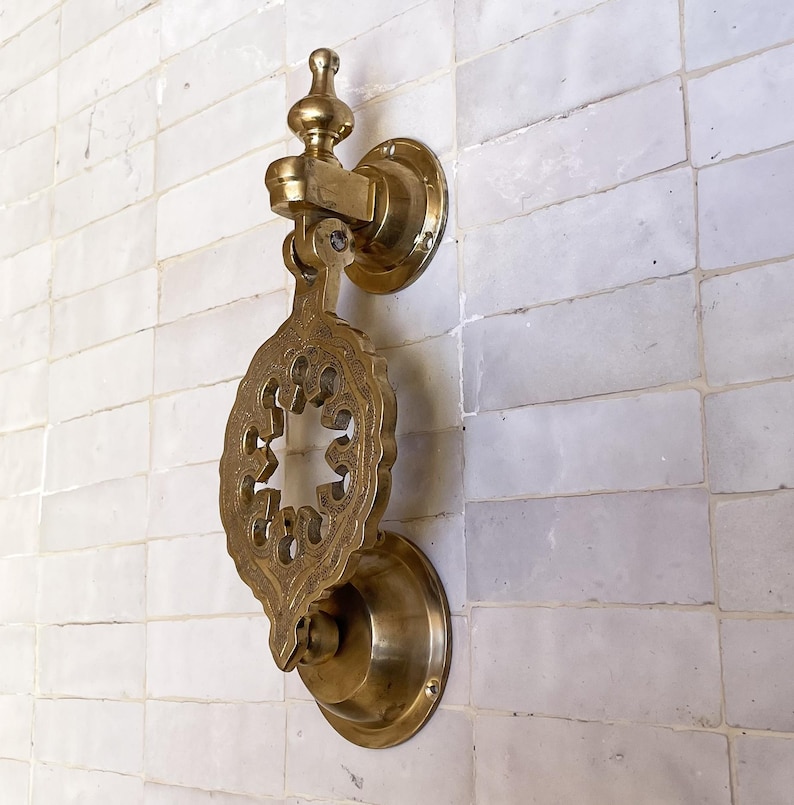 Puede incluir: Aldaba de puerta de lat&oacute;n dorado con un dise&ntilde;o calado decorativo. La aldaba est&aacute; montada en una pared de azulejos blancos. Tiene una base circular y una placa de golpeo detallada en forma de hoja. Acabado dorado antiguo.