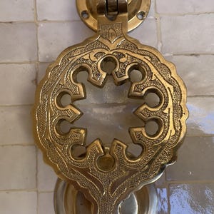 Puede incluir: Aldaba de puerta de lat&oacute;n ornamentada con un intrincado dise&ntilde;o calado. La aldaba presenta una forma central en forma de estrella rodeada por un borde detallado de inspiraci&oacute;n floral. El lat&oacute;n tiene una p&aacute;tina c&aacute;lida y envejecida, montada sobre una superficie de azulejos blancos.
