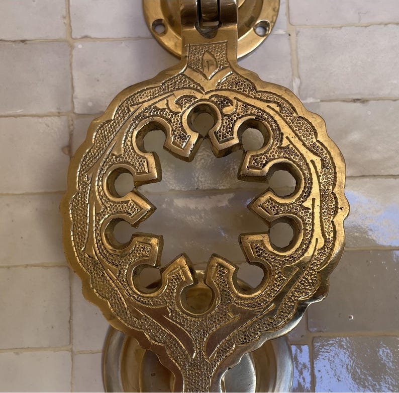 Puede incluir: Aldaba de puerta de lat&oacute;n ornamentada con un intrincado dise&ntilde;o calado. La aldaba presenta una forma central en forma de estrella rodeada por un borde detallado de inspiraci&oacute;n floral. El lat&oacute;n tiene una p&aacute;tina c&aacute;lida y envejecida, montada sobre una superficie de azulejos blancos.