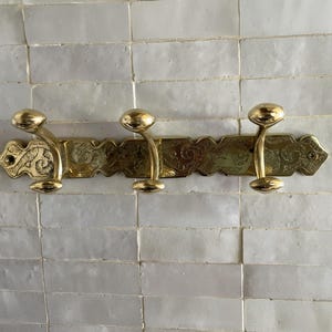 May include: A brass coat rack for wall mounting, featuring three curved hooks. The rack has an ornate, engraved design along the horizontal bar and decorative end pieces. The hooks have a rounded, knob-like shape.
