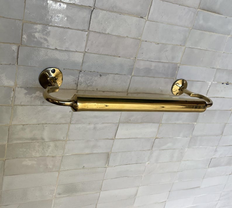 Peut inclure: Un porte-serviettes dor&eacute; avec une section centrale cylindrique et des extr&eacute;mit&eacute;s incurv&eacute;es. La barre est fix&eacute;e sur un mur carrel&eacute; de couleur claire. Le design est simple et &eacute;l&eacute;gant, adapt&eacute; &agrave; une salle de bain ou une cuisine. Mesure environ 40 cm de long.