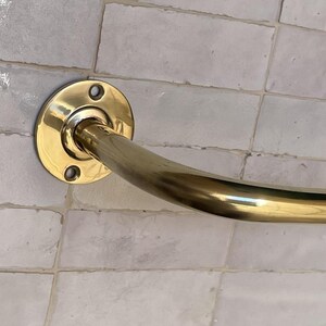 May include: A curved, brass-colored grab bar mounted on a light-colored tiled wall. The bar has a circular mounting plate with three screw holes. The curved section of the bar is smooth and shiny.