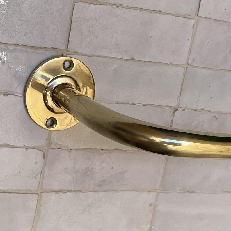 May include: A curved, brass-colored grab bar mounted on a light-colored tiled wall. The bar has a circular mounting plate with three screw holes. The curved section of the bar is smooth and shiny.