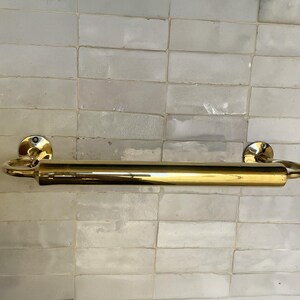 Peut inclure: Un porte-serviettes en laiton poli. La barre est fix&eacute;e sur un mur avec un motif de carreaux rectangulaires. La barre a un design cylindrique avec des extr&eacute;mit&eacute;s arrondies et des plaques de montage circulaires.