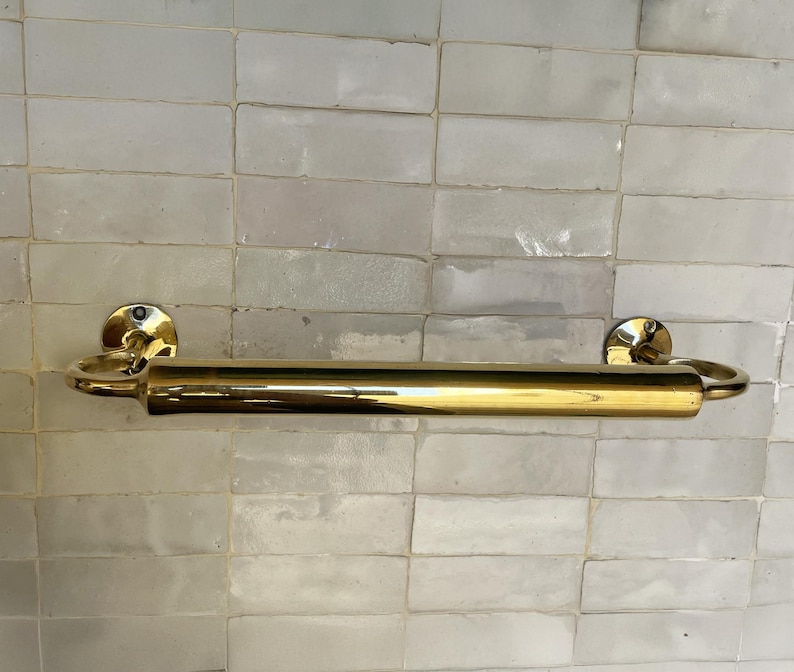 Peut inclure: Un porte-serviettes en laiton poli. La barre est fix&eacute;e sur un mur avec un motif de carreaux rectangulaires. La barre a un design cylindrique avec des extr&eacute;mit&eacute;s arrondies et des plaques de montage circulaires.
