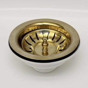 Puede incluir: Un colador de fregadero de latón y dorado con base blanca. El colador tiene un diseño circular con una serie de ranuras y un poste central. El borde es dorado y la base es blanca con un acento negro.