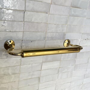 Peut inclure: Un porte-serviettes en laiton poli avec des bras incurv&eacute;s et des fixations murales circulaires. La barre est mont&eacute;e sur un mur carrel&eacute; blanc. Le laiton a une surface brillante et r&eacute;fl&eacute;chissante. La barre mesure environ 46 cm de long.