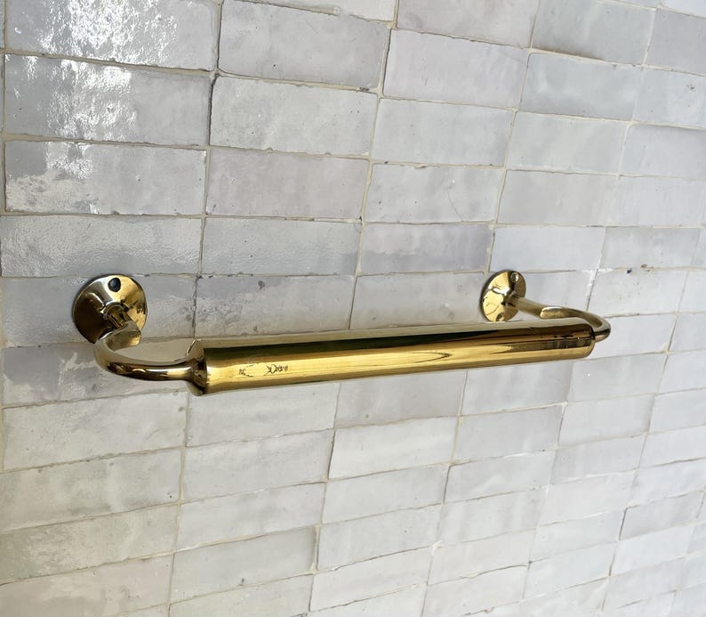 Peut inclure: Un porte-serviettes en laiton poli avec des bras incurv&eacute;s et des fixations murales circulaires. La barre est mont&eacute;e sur un mur carrel&eacute; blanc. Le laiton a une surface brillante et r&eacute;fl&eacute;chissante. La barre mesure environ 46 cm de long.