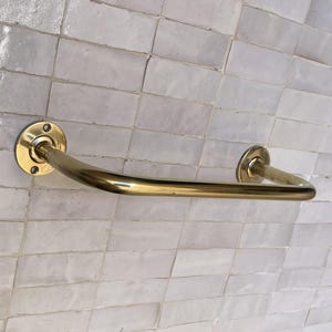 May include: A brass-colored towel bar with a curved design. The bar is mounted on a white tiled wall. The bar has a circular mounting plate on each end with screw holes.