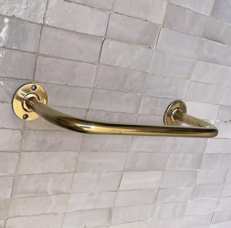 May include: A brass-colored towel bar with a curved design. The bar is mounted on a white tiled wall. The bar has a circular mounting plate on each end with screw holes.
