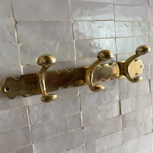 Peut inclure: Un porte-manteau mural en laiton avec trois crochets incurvés. Le porte-manteau a un bord décoratif festonné et est monté sur un mur carrelé blanc. Les crochets sont de forme arrondie, en forme de champignon.