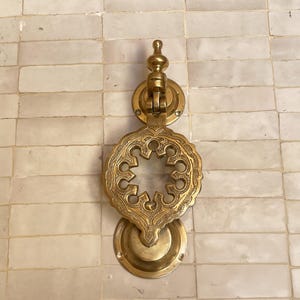 Puede incluir: Aldaba de puerta de lat&oacute;n dorado con un dise&ntilde;o calado intrincado. La aldaba presenta una parte superior circular, un motivo central floral y una base redondeada. El lat&oacute;n tiene un tono dorado c&aacute;lido y est&aacute; montado sobre una superficie clara.