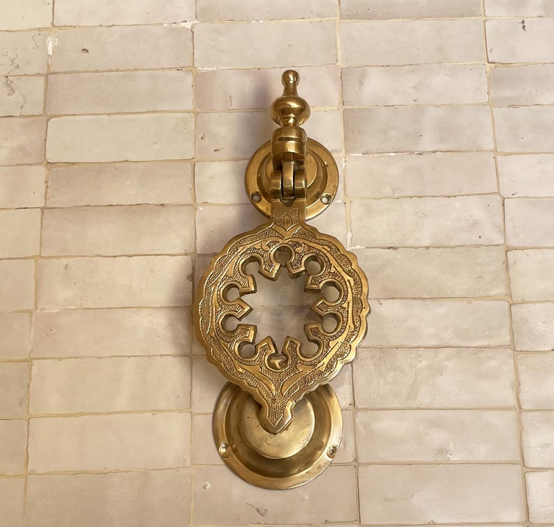 Puede incluir: Aldaba de puerta de lat&oacute;n dorado con un dise&ntilde;o calado intrincado. La aldaba presenta una parte superior circular, un motivo central floral y una base redondeada. El lat&oacute;n tiene un tono dorado c&aacute;lido y est&aacute; montado sobre una superficie clara.