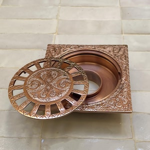 May include: An ornate copper-coloured drain cover. The square frame and circular cover feature detailed relief designs. The cover is partially open, revealing the drain's interior. The design is intricate and artistic, with swirling patterns. The drain is likely for a shower or bathroom.