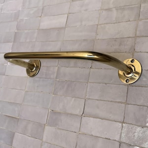 May include: A curved, brass-colored grab bar mounted on a light gray tiled wall. The bar has a smooth, polished finish and is attached to the wall with circular mounting plates. The design is simple and functional.