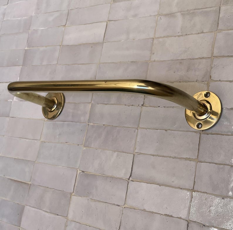 May include: A curved, brass-colored grab bar mounted on a light gray tiled wall. The bar has a smooth, polished finish and is attached to the wall with circular mounting plates. The design is simple and functional.