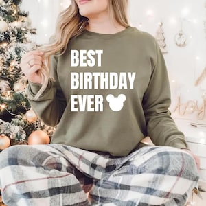 May include: An olive green sweatshirt with the text "BEST BIRTHDAY EVER" in white, along with a white Mickey Mouse head graphic. The person is wearing plaid pajama pants. The background includes a Christmas tree and decorative lights.