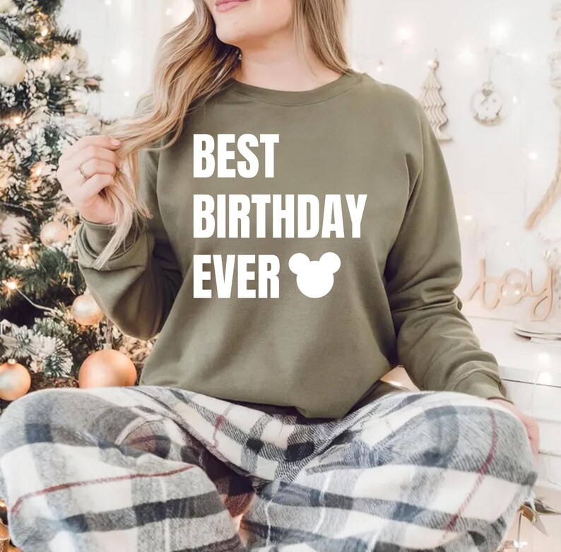 May include: An olive green sweatshirt with the text "BEST BIRTHDAY EVER" in white, along with a white Mickey Mouse head graphic. The person is wearing plaid pajama pants. The background includes a Christmas tree and decorative lights.