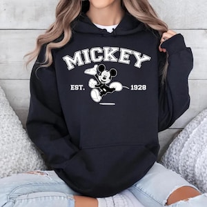 May include: Black hooded sweatshirt featuring the word "MICKEY" in white block letters above a graphic of Mickey Mouse. Below the graphic are the words "EST. 1928". The sweatshirt is a dark color and has a drawstring hood.
