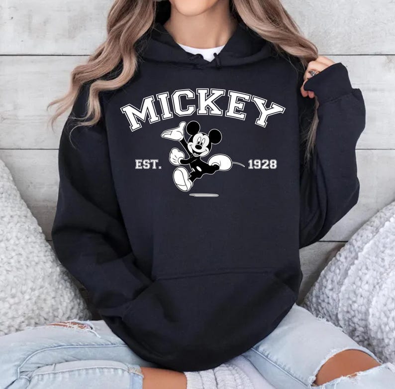 May include: Black hooded sweatshirt featuring the word "MICKEY" in white block letters above a graphic of Mickey Mouse. Below the graphic are the words "EST. 1928". The sweatshirt is a dark color and has a drawstring hood.