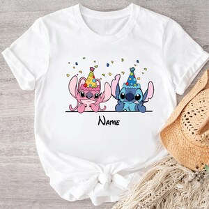 May include: White t-shirt with a pink and blue Stitch character wearing party hats. Confetti is scattered around the characters. The text "Name" is printed below the characters.