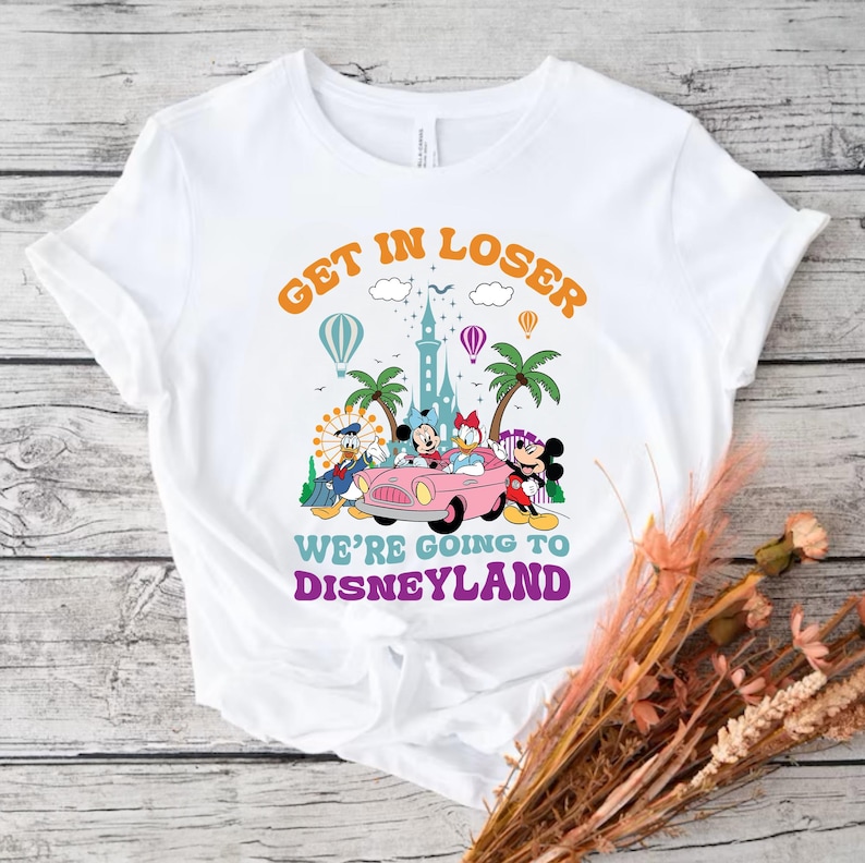 May include: White t-shirt with a graphic of Mickey Mouse and friends driving a pink car with the text "Get in Loser We're Going to Disneyland" in a retro font.