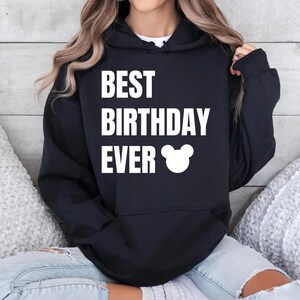 May include: A black hooded sweatshirt with the text "BEST BIRTHDAY EVER" in white block letters. A white Mickey Mouse head graphic is below the text. The sweatshirt is a casual style.