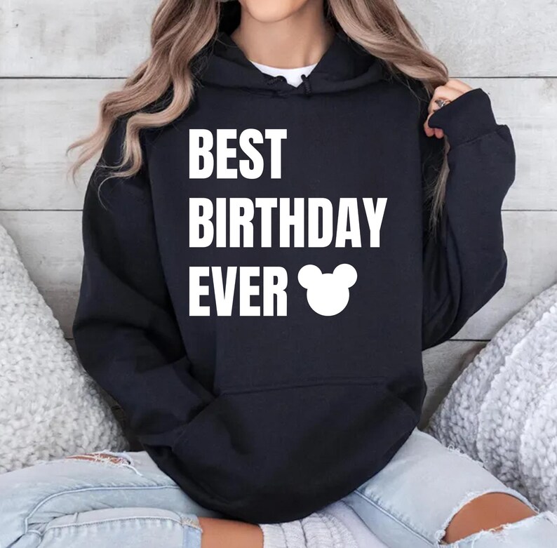 May include: A black hooded sweatshirt with the text "BEST BIRTHDAY EVER" in white block letters. A white Mickey Mouse head graphic is below the text. The sweatshirt is a casual style.