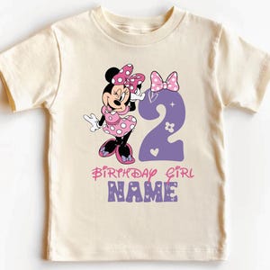 Może przedstawiać: Jasnobeżowa koszulka z postacią Minnie Mouse w r&oacute;żowe i białe kropki trzymającą fioletową dw&oacute;jkę. Na koszulce widnieje napis "Birthday Girl" i "NAME" w kolorze r&oacute;żowym i fioletowym.