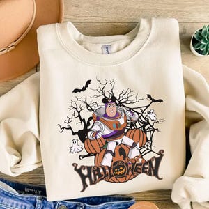 K&ouml;nnte beinhalten: Cremefarbenes Sweatshirt mit einem Halloween-Motiv. Das Design zeigt eine Zeichentrickfigur in einem Raumanzug, K&uuml;rbisse, Flederm&auml;use, Geister und das Wort "HALLOWEEN". Die Grafik ist in Orange, Schwarz, Wei&szlig; und Lila gehalten.